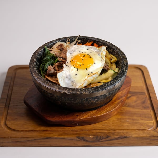 Bibimbap in a stone bowl with a fried egg, beef, and vegetables on a wooden board.