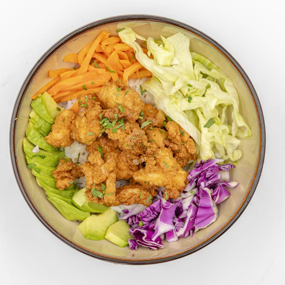 Bowl of crispy fried chicken over rice with lettuce, carrots, avocado, cucumber, and purple cabbage.