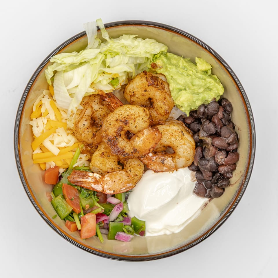 Top-down view of a shrimp burrito bowl with rice, beans, lettuce, cheese, guacamole, sour cream, and fresh salsa.
