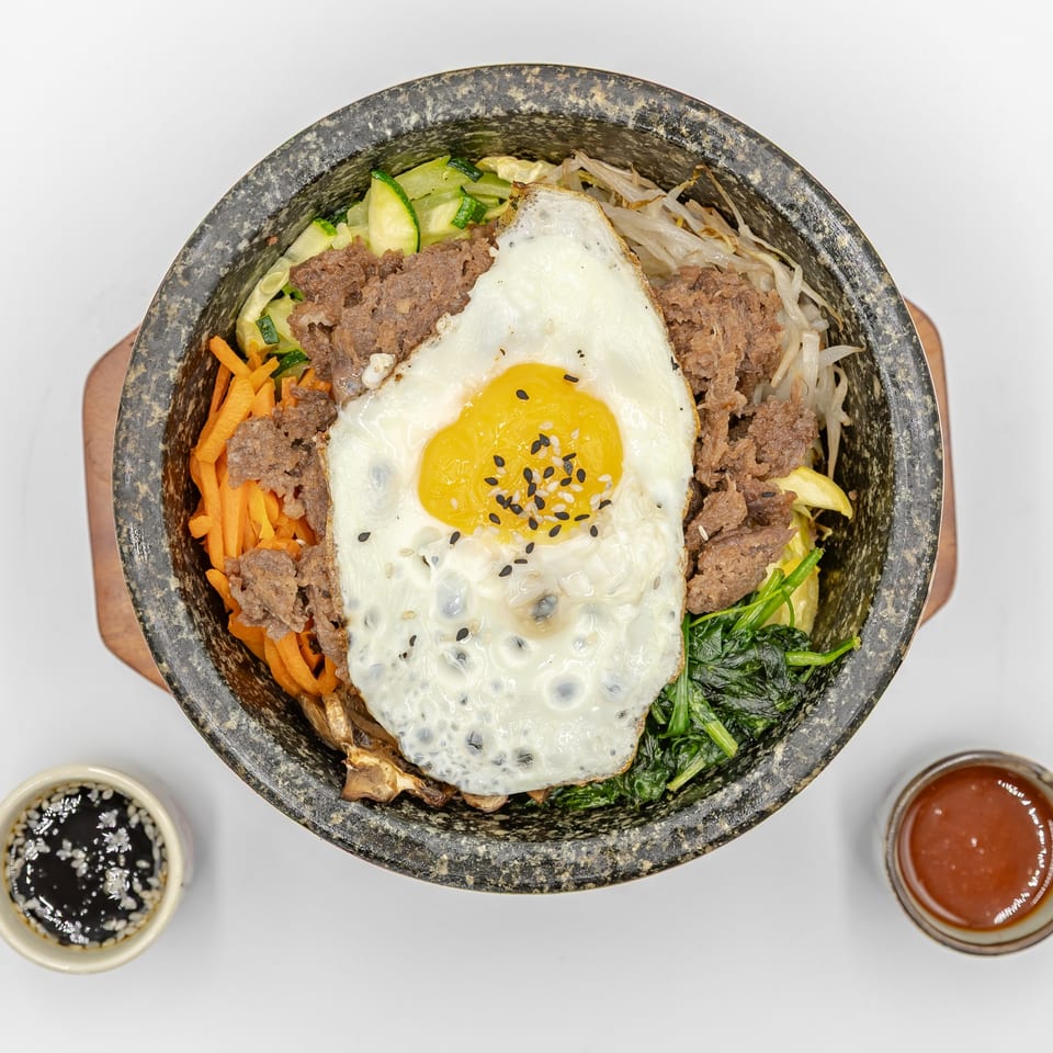 Korean bibimbap in a stone bowl topped with a fried egg, beef, vegetables, and sesame seeds, with sauces alongside.